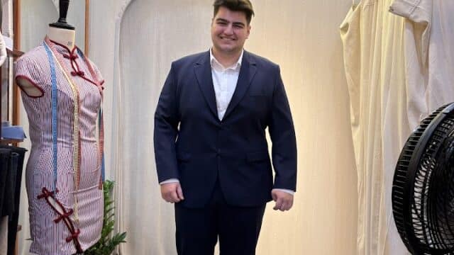 A man in a navy suit stands barefoot in a tailor shop, smiling under a neon Van's Tailor sign showcasing custom tailored suits.