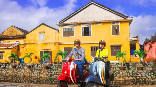 Two people on scooters enjoy a sunny day in front of colorful yellow buildings, perfect for Vespa Adventures and memorable scooter tours.