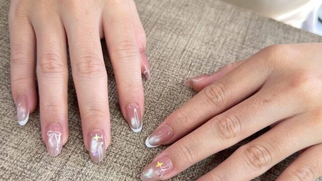 Hands with almond-shaped, nude nails from Vie Spa and Nail feature white tips with delicate ribbon and star nail art designs.