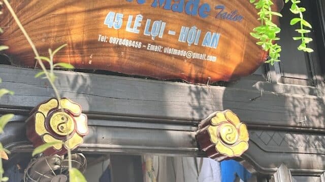 Wooden sign reading Viet Made Tailor, 45 Le Loi - Hoi An above a shop entrance with clothes and lush greenery in Hoi An.