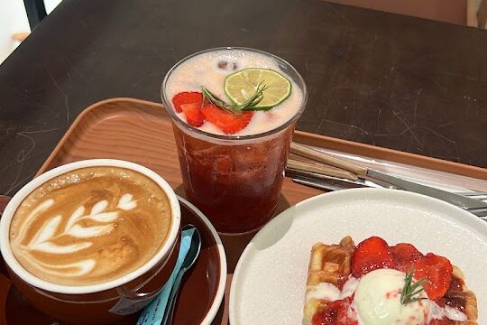 A latte, strawberry drink, and waffle with cream and strawberries on a tray at a café table.