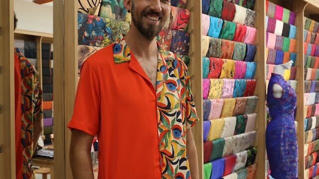 Man smiling in a Hoi An fabric shop, wearing a bright orange shirt with a colorful patterned collar and pocket.