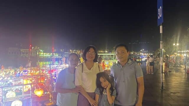A family of four poses at night by a brightly lit riverside, ready for a Lantern Boat Tour with colorful boats and lights in the background.