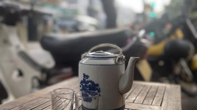 A teapot and two glasses sit on a wooden table outdoors in Hoi An, with motorbikes and trees in the background.