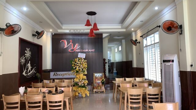 Small Hoi An restaurant interior with wooden chairs, flower stand, neon sign, and ceiling fans on the walls.