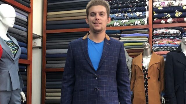 A man in a blue blazer stands in a Hoi An fabric shop, surrounded by mannequins and shelves with folded fabrics.