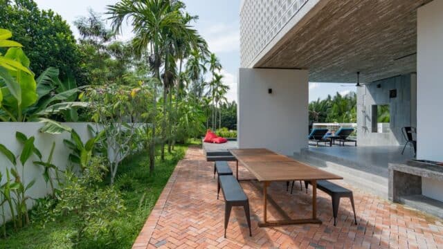 Modern patio with a wooden table, benches, lush greenery, and Hoi An-inspired lounge chairs in the open house area.