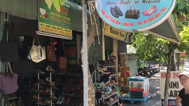 Shoe shop in Hoi An with a 447 sign, shoes and bags outside, and a woman by the entrance on a sunny day.