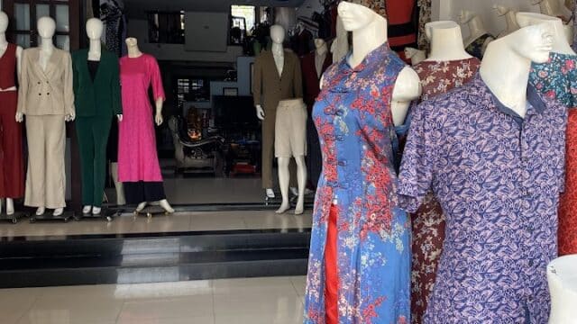 Mannequins in colorful dresses and suits are displayed inside and outside a Hoi An clothing store with tiled floors.