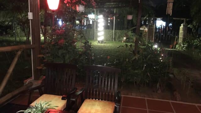 Outdoor patio at night evokes Hoi An charm, with two wooden chairs, a table with a plant, red lantern, and glowing garden lights.