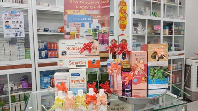 A Hoi An pharmacy counter displays skincare, hand sanitizers, and lucky gift boxes; medicine shelves are visible behind.