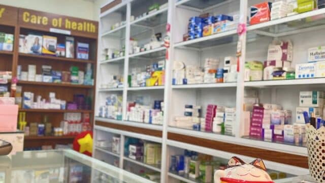 A pharmacy interior in Hoi An with shelves of medicines, a Maneki-neko cat figurine, and business cards on the counter.