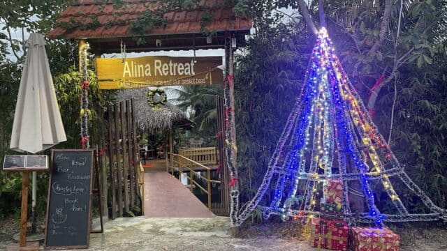 Entrance to Alina Retreat in Hoi An with a yellow sign, menu board, and a Christmas tree of lights surrounded by wrapped gifts.