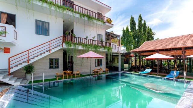 Modern Hoi An guesthouse with balconies, a pool, lounge chairs, and umbrellas under a sunny blue sky.