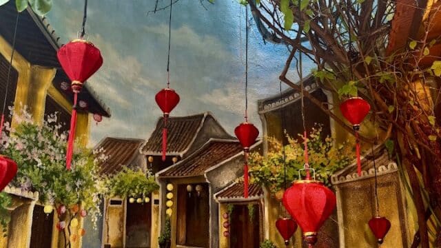 A mural of a curved street in Hoi An with yellow houses, red lanterns hanging, and green vines above.