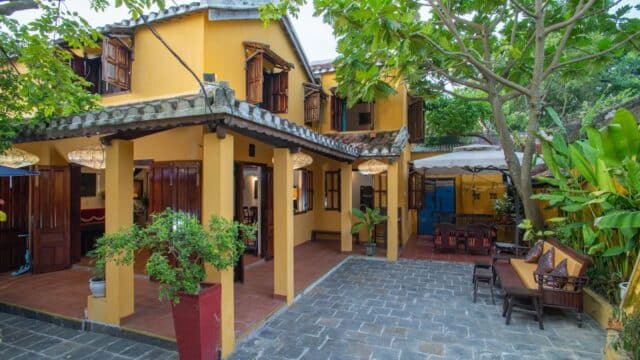 A yellow two-story Hoi An house with wooden shutters, patio seating, and lush greenery in the courtyard.