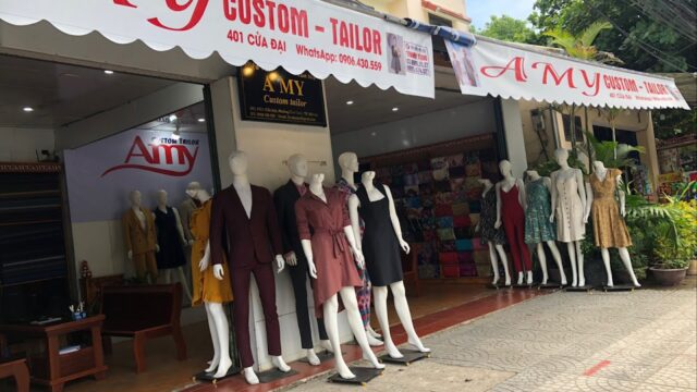 Mannequins in vibrant outfits stand outside AMY, a Hoi An tailor shop, with colorful fabrics displayed by the entrance.