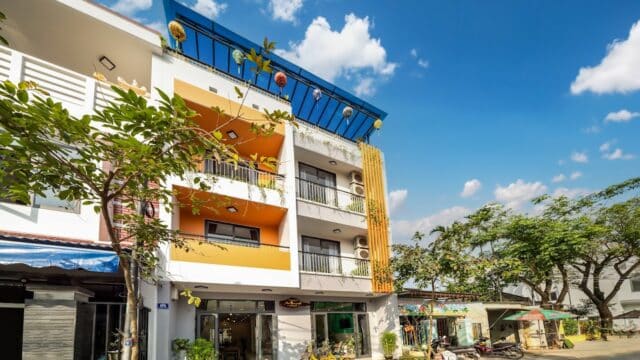 Modern three-story building with balconies and bright yellow accents in Hoi An, set among trees under a blue sky with clouds.