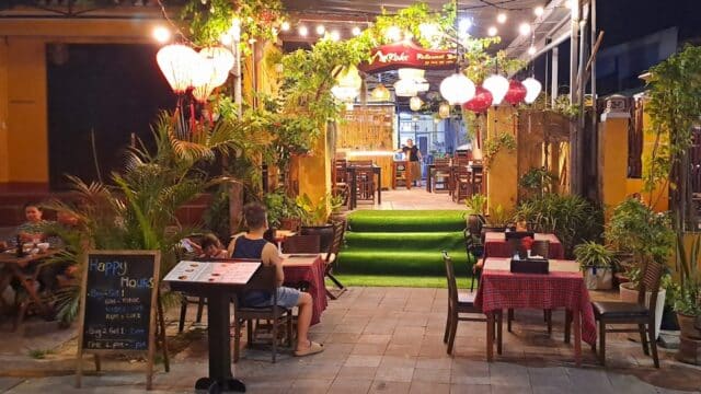 Outdoor restaurant in Hoi An with string lights, lanterns, and people dining at tables on a patio surrounded by greenery.