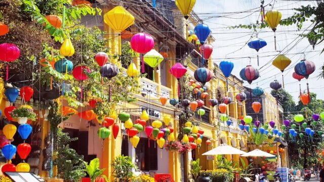 Colorful lanterns hang over a lively Hoi An street with yellow buildings, potted plants, and people in the background.