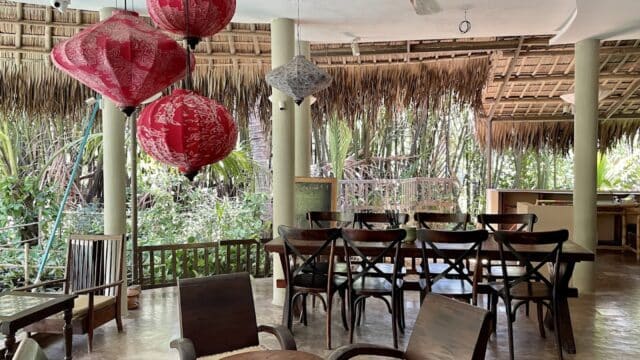 Open-air Hoi An dining area with wooden chairs, red lanterns, and thatched roof, surrounded by lush green tropical plants.