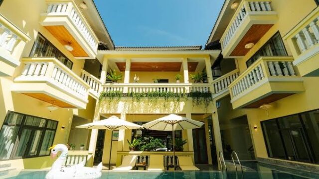 A sunny pool area with a swan float and umbrellas, surrounded by a yellow, modern three-story Hoi An building.