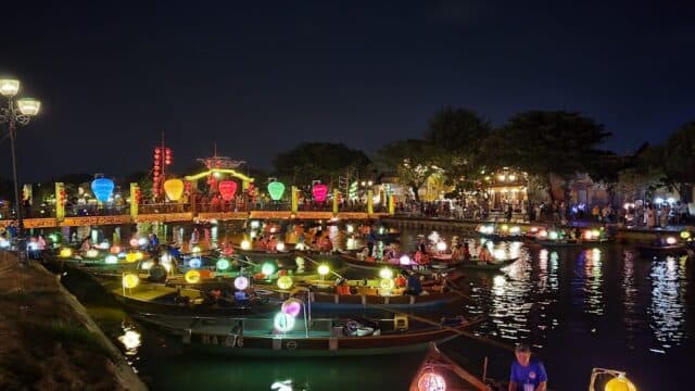 Colorful lanterns light up boats on Hoi An’s river at night, with crowds and illuminated buildings glowing in the background.