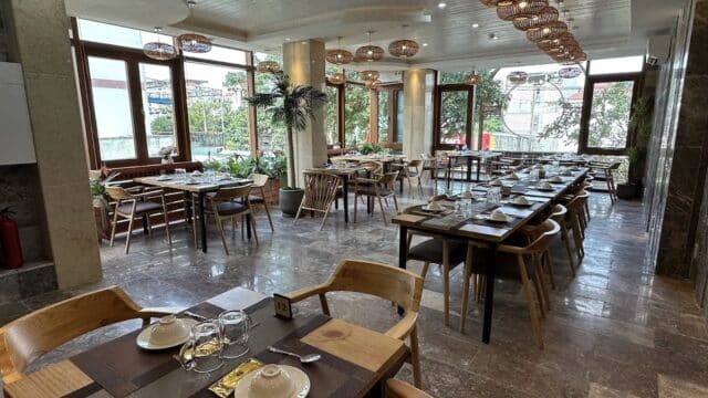 A spacious, well-lit Hoi An restaurant with wooden tables and chairs, large windows framing greenery, and tables set for dining.