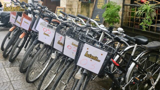 Row of rental bicycles with numbered labels parked on a Hoi An sidewalk near a stone building and signboards.