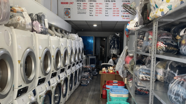 A Hoi An laundromat with washing machines left, laundry baskets and folded clothes right, and prices displayed above.