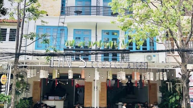 A modern café in Hoi An with outdoor seating, blue windows, and green trees in front on a sunny day.