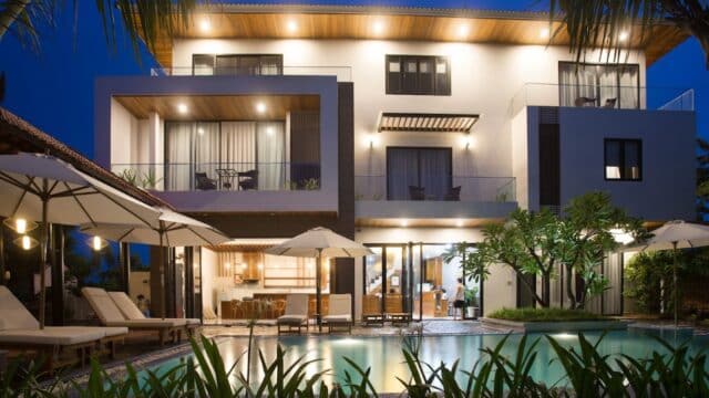 Modern three-story villa in Hoi An with large windows, balconies, poolside lounge chairs, umbrellas, and palm trees, illuminated at night.