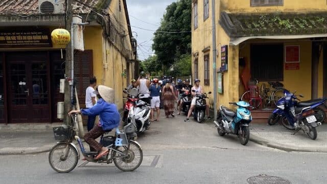 A person rides a bicycle past yellow Hoi An buildings and parked motorbikes on a busy street.