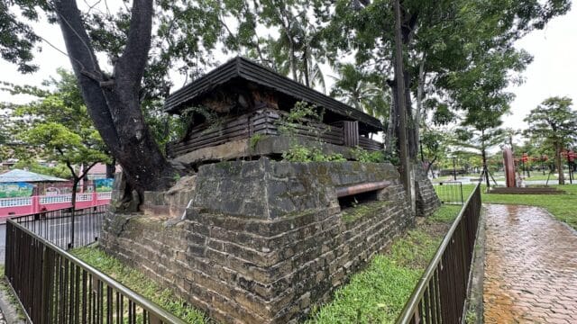 An old stone structure with a wooden roof in Hoi An, fenced off in a park with trees and a brick walkway.
