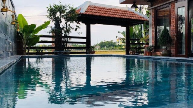 A clear swimming pool beside a brick house with balconies, a red-tiled gazebo, and Hoi An charm in the background.