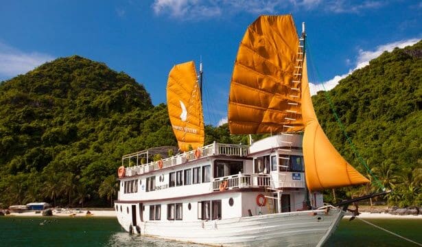 A white boat with orange sails floats near a lush, green island under a blue Hoi An sky.