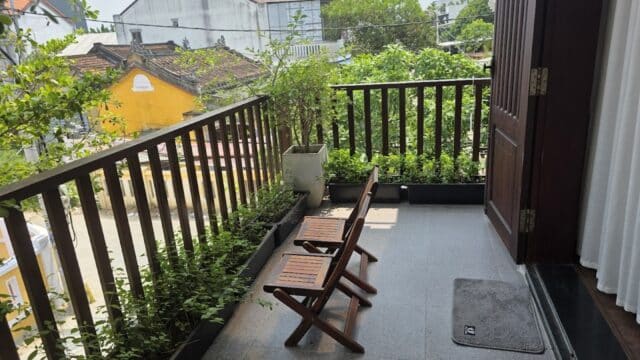 Two wooden chairs on a small balcony with potted plants, overlooking a quiet Hoi An street lined with houses and lush greenery.