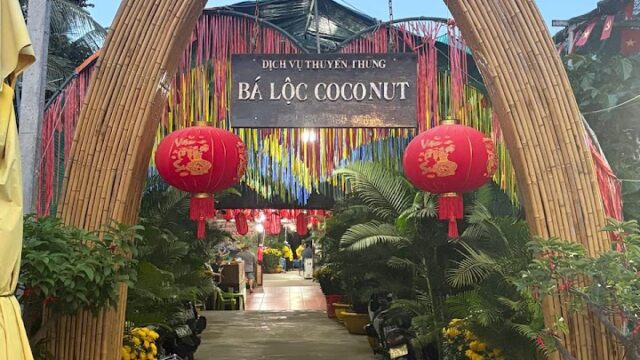 Entrance with bamboo arch, red lanterns, and a Bá Lộc Coconut sign evokes Hoi An vibes, adorned with streamers and potted plants.