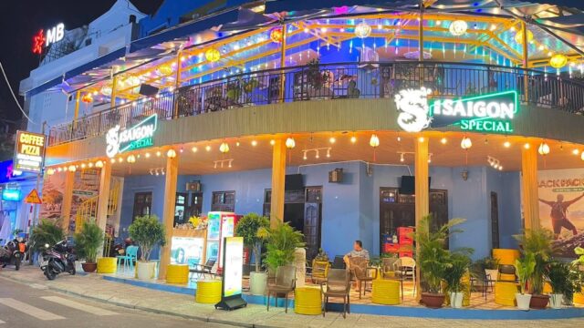 A brightly lit café with Saigon Special signs, outdoor seating, plants, and neon lights evokes the vibrant charm of Hoi An at night.