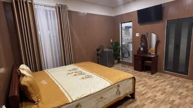 Neatly made bed in a tidy Hoi An bedroom with wood paneling, a vanity, wardrobe, TV, lamp, and curtains.