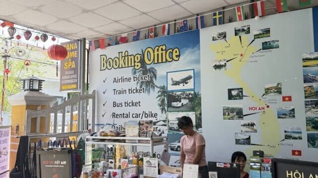 Two people work at a booking office desk in Hoi An, with travel posters, flags, and signs for tickets and rentals behind them.