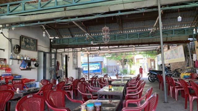 An open-air eatery in Hoi An with empty red plastic chairs and tables, drink shelves, and a motorbike parked outside.