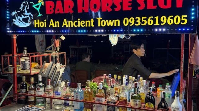 A street bar cart in Hoi An with a neon BAR HORSE SLUT sign, bottles of alcohol, and menus on display.