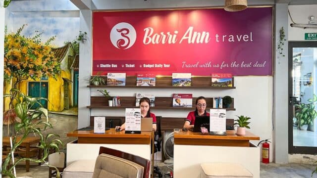 Two women sit at desks in a bright travel agency office, with a red Barri Ann Travel sign and a map of Hoi An behind them.