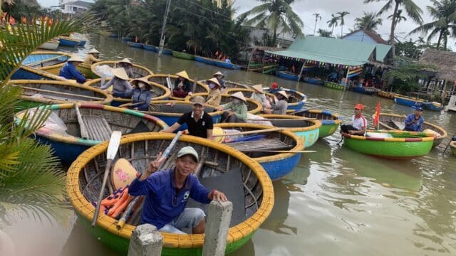 People in round basket boats on a river in Hoi An, wearing hats, with palm trees and buildings in the background.