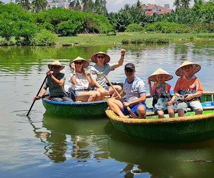 Five people in round boats on a river in Hoi An, wearing traditional hats, surrounded by lush greenery and blue sky.