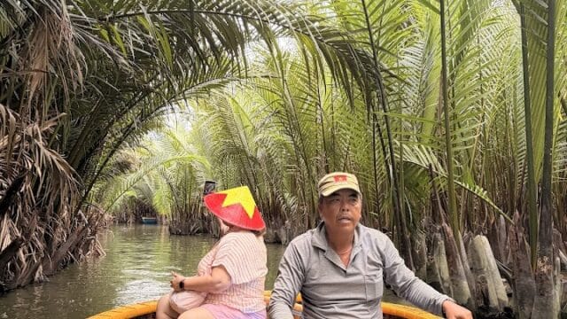A man and child in a round boat paddle through lush green palm trees on a narrow waterway near Hoi An.