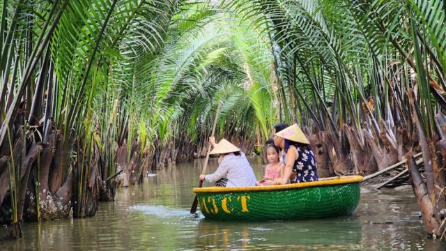 Three people in a round boat paddle through Hoi An’s waterways, surrounded by lush palms and wearing traditional conical hats.