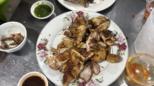 Grilled meats on floral Hoi An plates, dipping sauces, greens, and drinks on a metal table; a hand holds a phone in the corner.