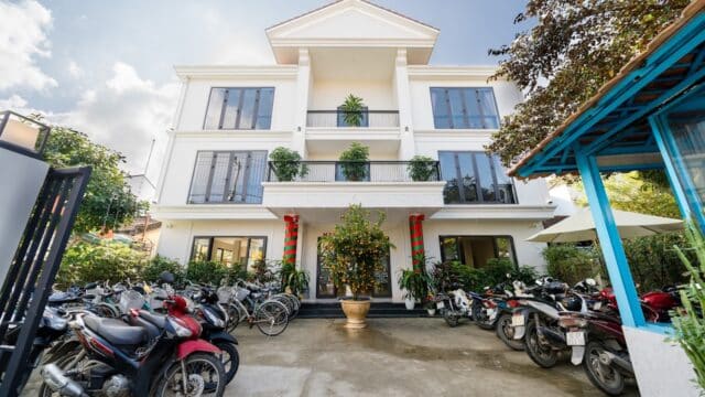 A white three-story building with balconies, parked motorbikes, and plants at the entrance in sunny Hoi An.